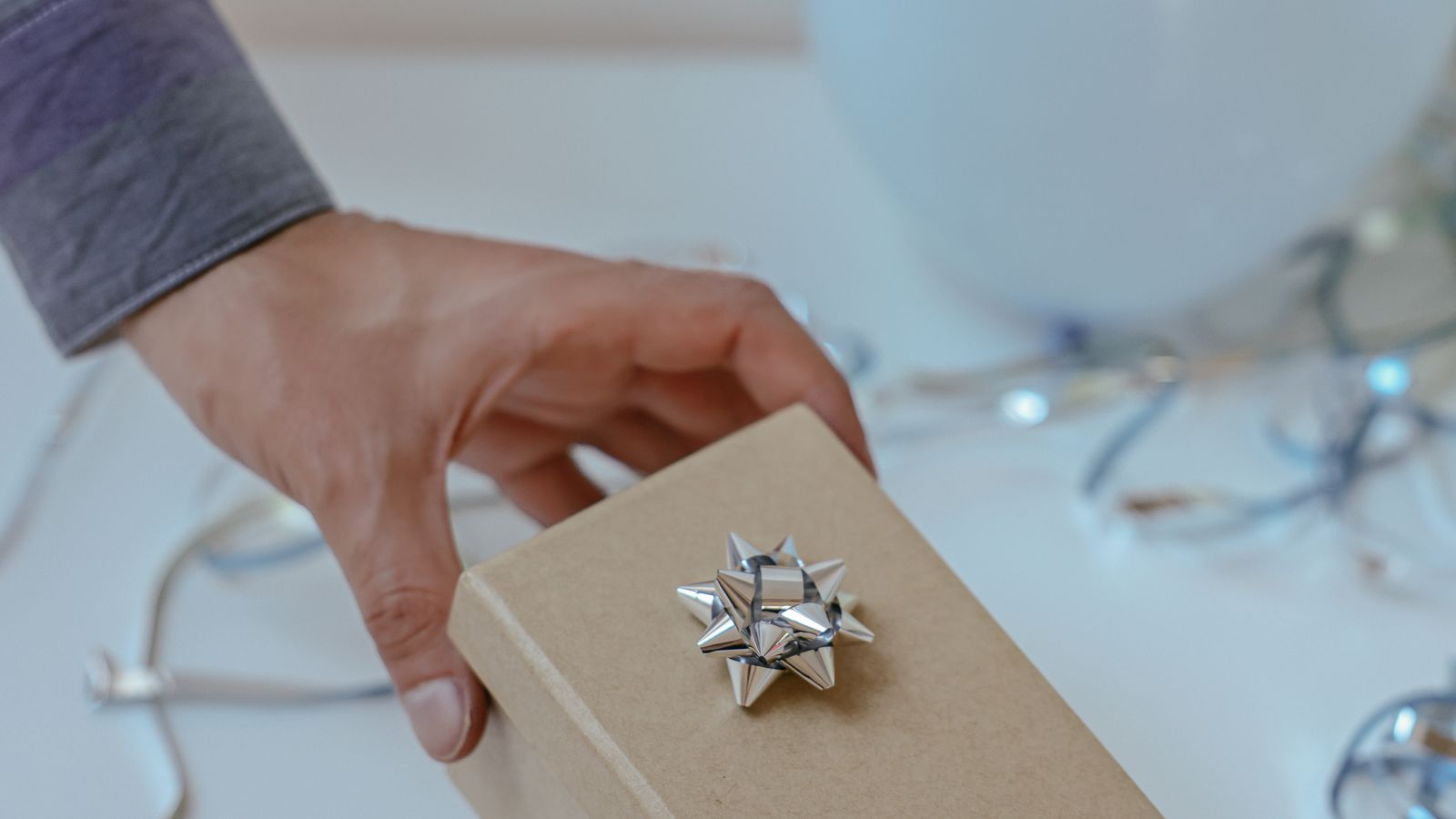 A hand reaching for a kraft paper gift box with a silver bow amid curled ribbon, last minute birthday gift UK